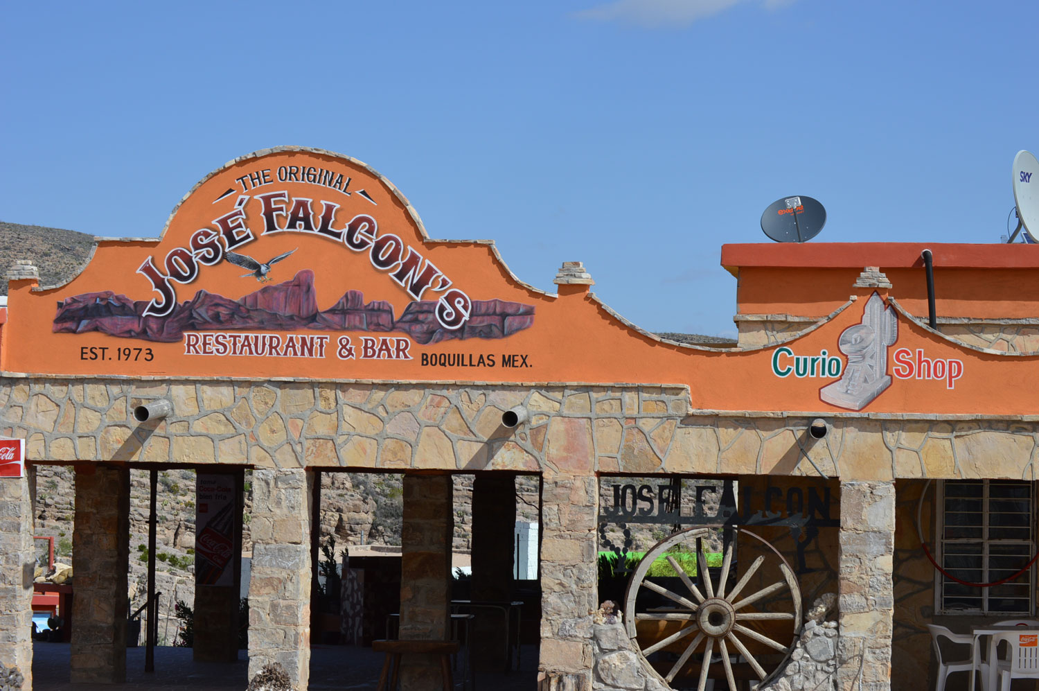 Front of José Falcon Restaurant in Boquillas, Mexico, with a sign painted mostly orange background and scenes of the Sierra del Carmen mountains. Building is made from rock with old wagon wheel decor mounted between porch pillars
